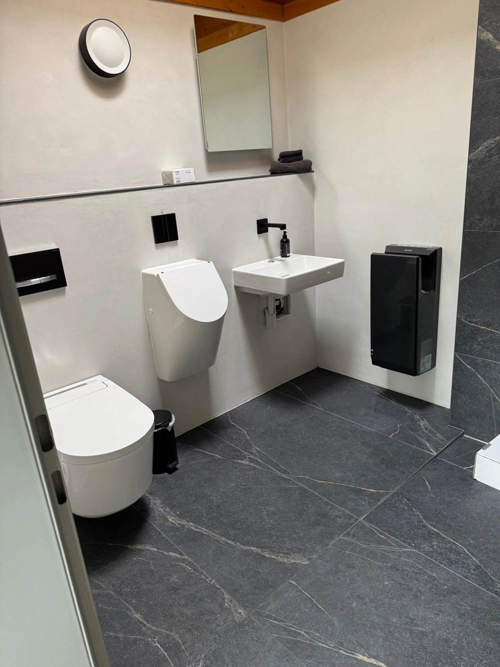 Moderne Badezimmer mit Toilette, Urinal, Wandwaschbecken und dunklem Bodenbelag nach Badsanierung