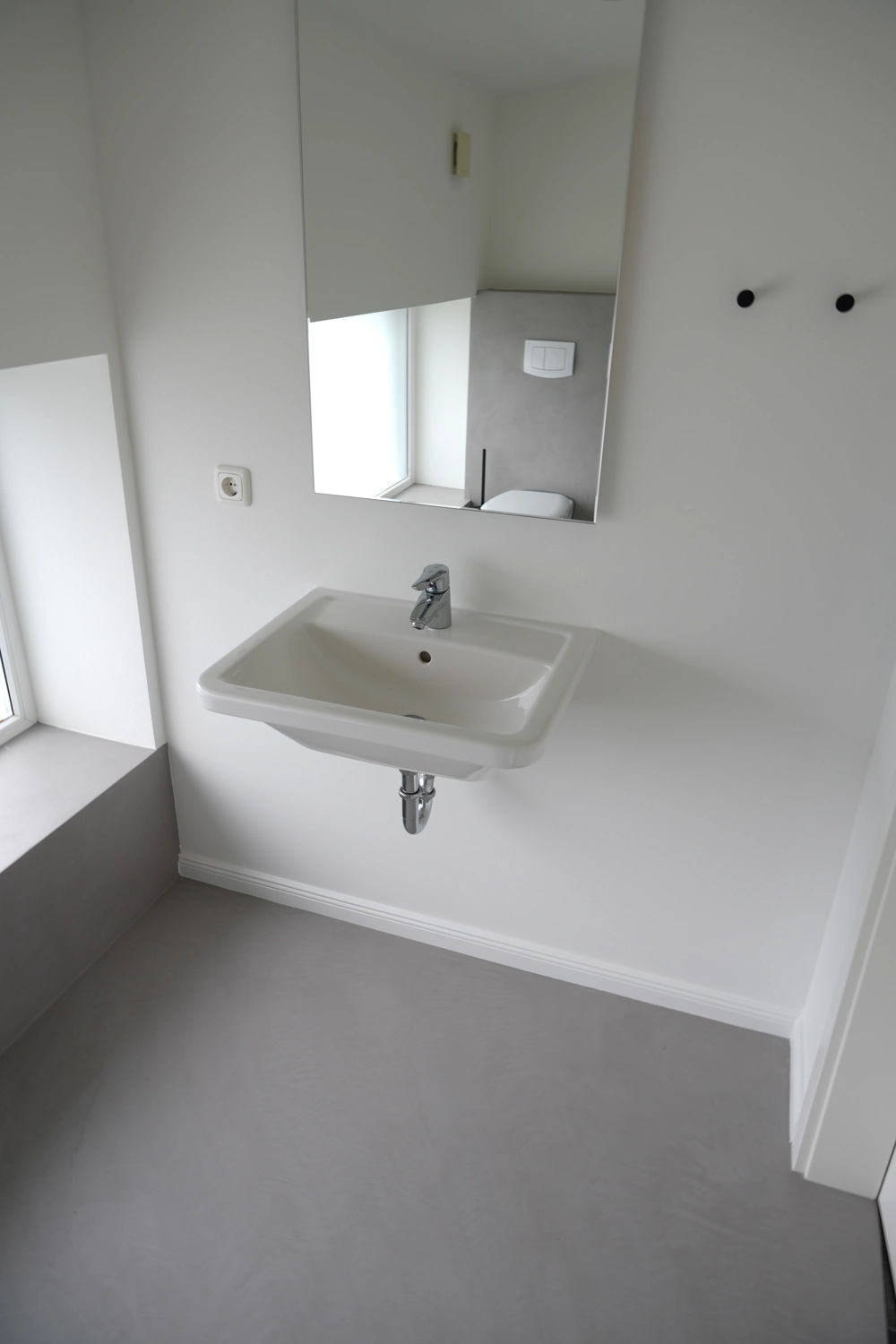 Helle Badsanierung mit wandhängendem Waschbecken, großem Spiegel und glattem grauen Bodenbelag