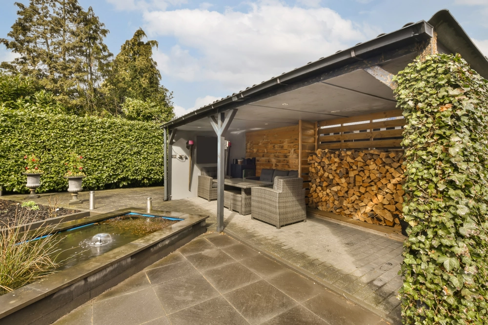 Terrassenüberdachung aus Holz mit Sitzbereich, Kaminholzlager und angrenzendem Teich im Garten