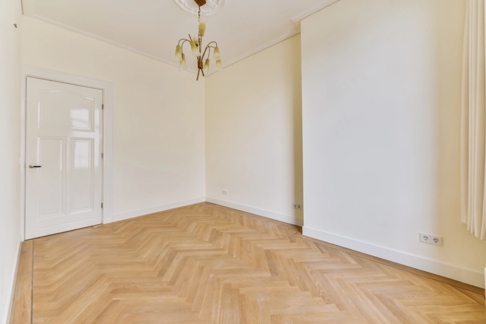 Leerraum mit weißen Wänden, Deckenlampe und frisch verlegtem Fischgrätparkett (Parkett verlegen)