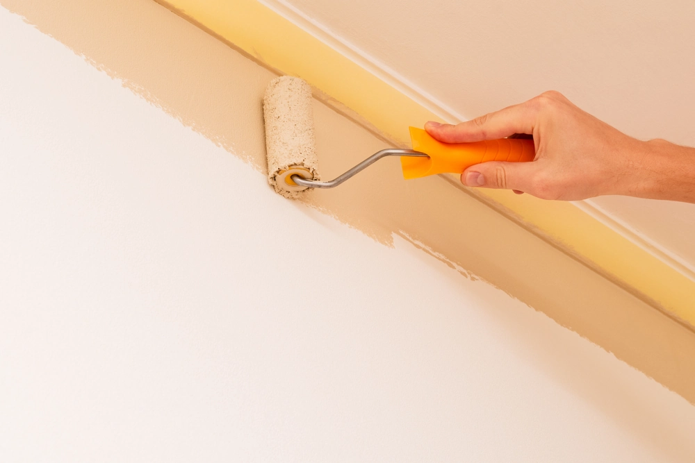 Hand mit Farbroller streicht beige Wandkante an der Decke, Malerarbeiten