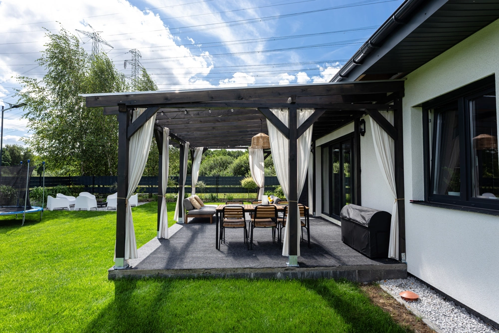 Pergola mit weißem Vorhang und Essbereich auf gepflasterter Terrasse neben Haus und Rasen